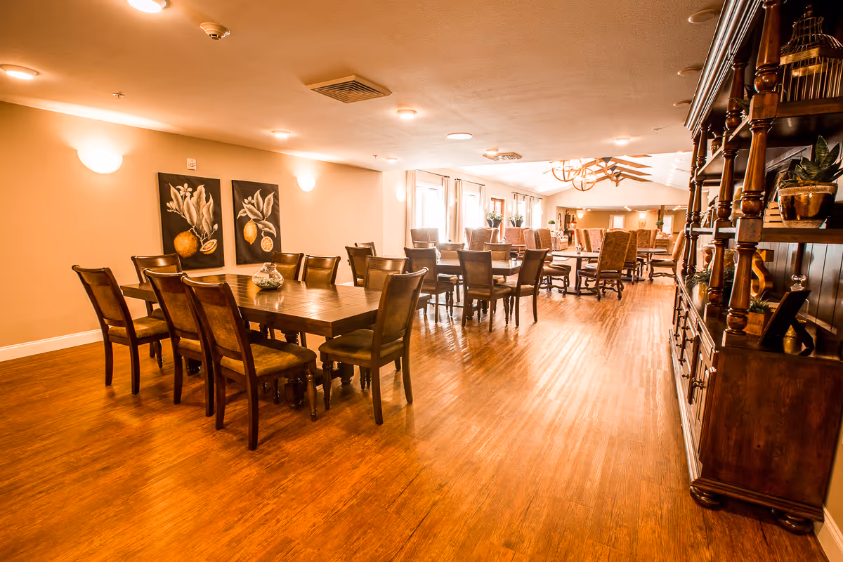 A spacious dining area with multiple wooden tables and chairs arranged neatly on a polished wooden floor. The room is warmly lit with wall sconces and ceiling lights, and decorated with framed fruit artwork on the wall. Large windows along one side let in natural light, and a wooden cabinet with decorative items is visible on the right.