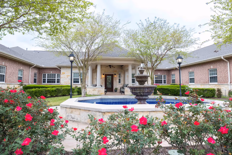 Courtyard view of a brick senior living facility showing a central tiered fountain, flowering bushes, lamp posts, and a covered columned entrance.