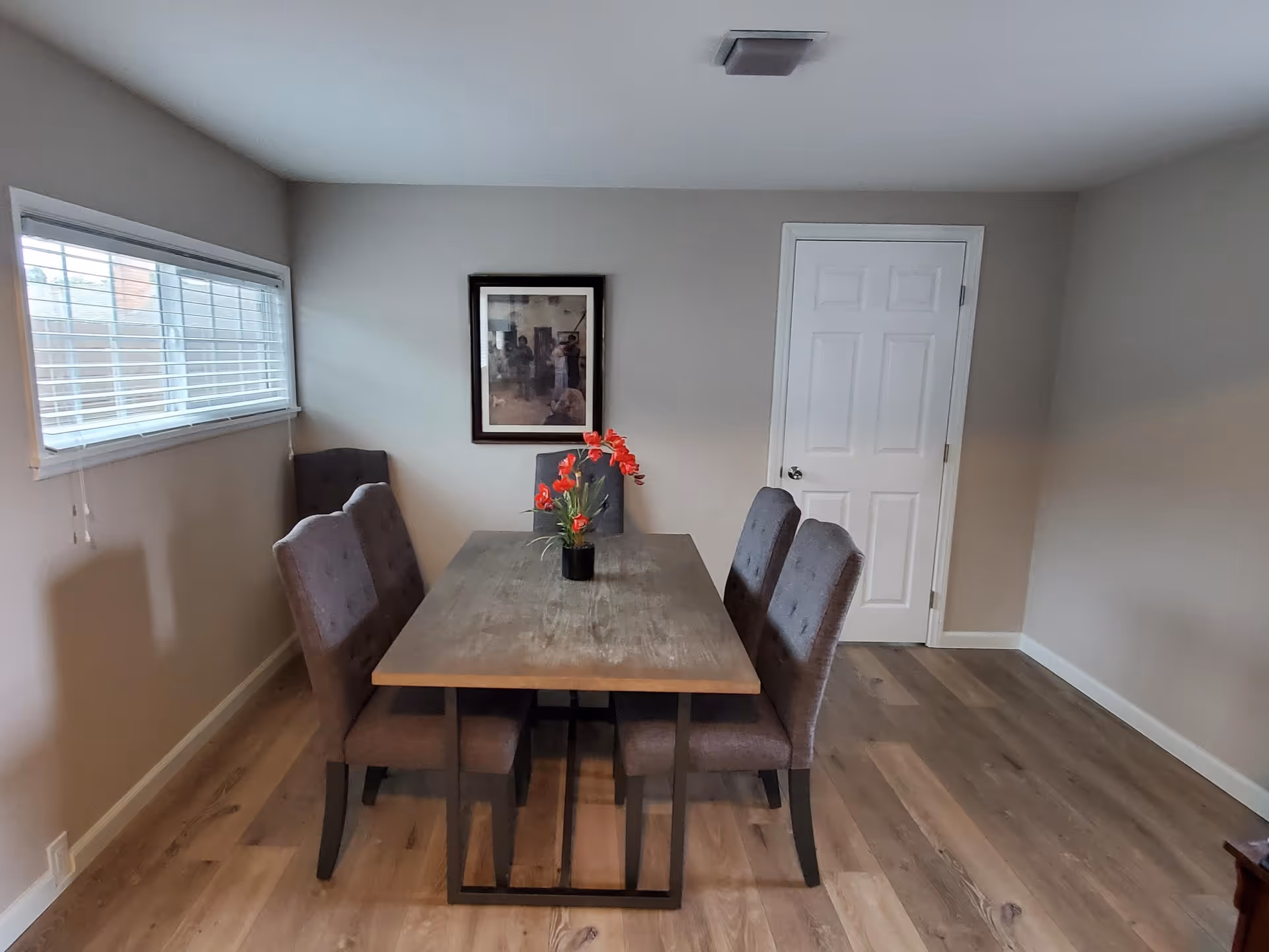 A dining room with a rectangular wooden table surrounded by six upholstered chairs. A vase with red flowers is placed in the center of the table. There is a window with blinds on the left wall and a framed picture hanging on the wall behind the table. A closed white door is visible on the right side of the back wall. The floor is wooden and the walls are painted light gray.