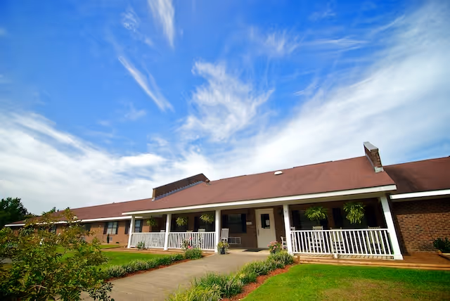 Exterior view of a single-story brick assisted living facility with a red roof, white railings on the porch, hanging plants, and a well-maintained lawn under a blue sky with wispy clouds.