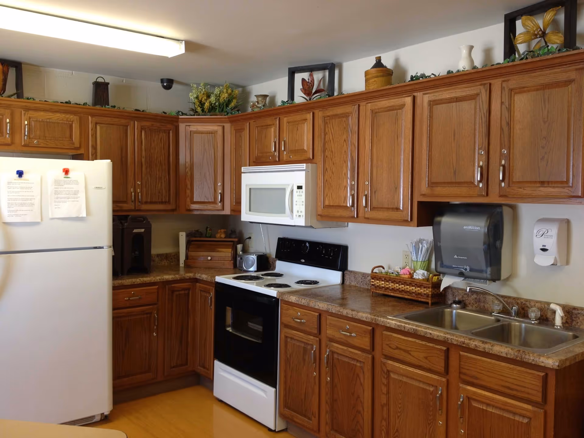 A kitchen with wooden cabinets, a white refrigerator with papers attached by magnets, a white microwave above a black and white electric stove, a double sink with a paper towel dispenser and soap dispenser on the wall, and various decorative items on top of the cabinets and countertop.