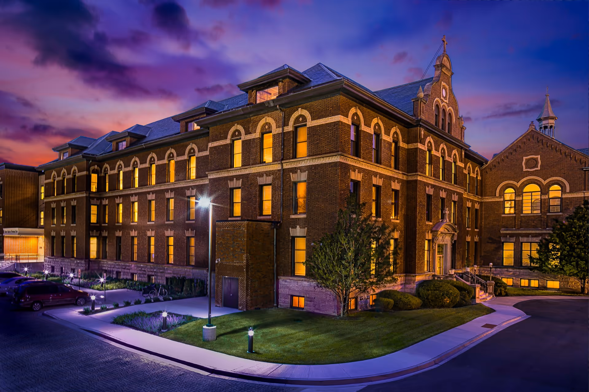 Exterior view of a large, historic brick building at dusk with illuminated windows and a well-maintained lawn and sidewalk in front. The sky is a vibrant mix of purple and orange hues.