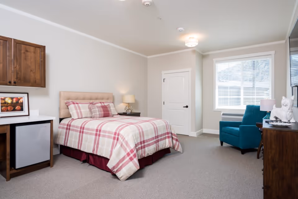 A senior living facility bedroom with a bed dressed in a red and beige plaid comforter and pillows, a wooden nightstand with a lamp, a teal armchair near a window with white blinds, a wooden dresser with a white dog figurine and a lamp, and a small refrigerator under a wooden cabinet.