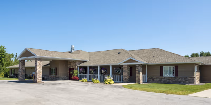 Single-story senior living building with a covered entrance supported by stone pillars, a front porch with hanging plants, lawn, and a paved driveway under a clear blue sky.