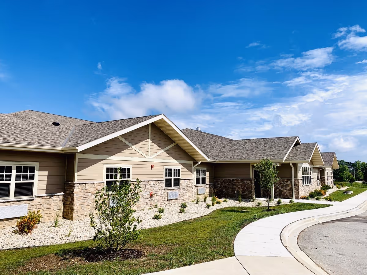 Front exterior of a single-story assisted living building with stone and siding, landscaped grounds, a curved sidewalk, and blue sky.