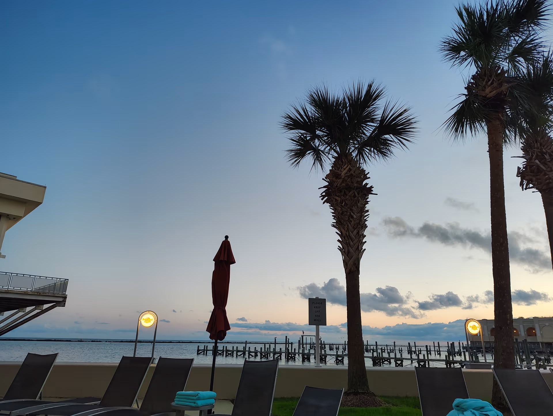 Sunset view of a waterfront outdoor lounge area with palm trees and chairs.