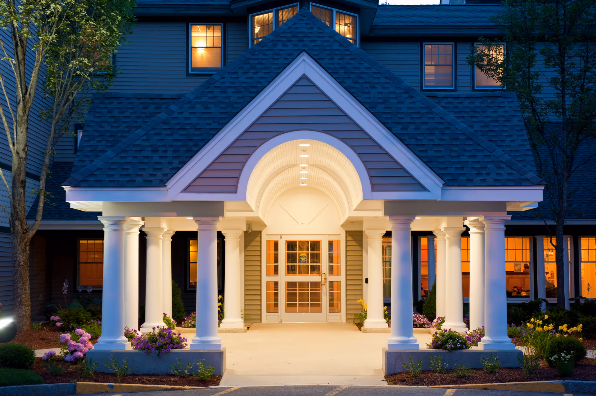 Columned portico entrance of a residential building with an illuminated doorway and landscaped beds at dusk.