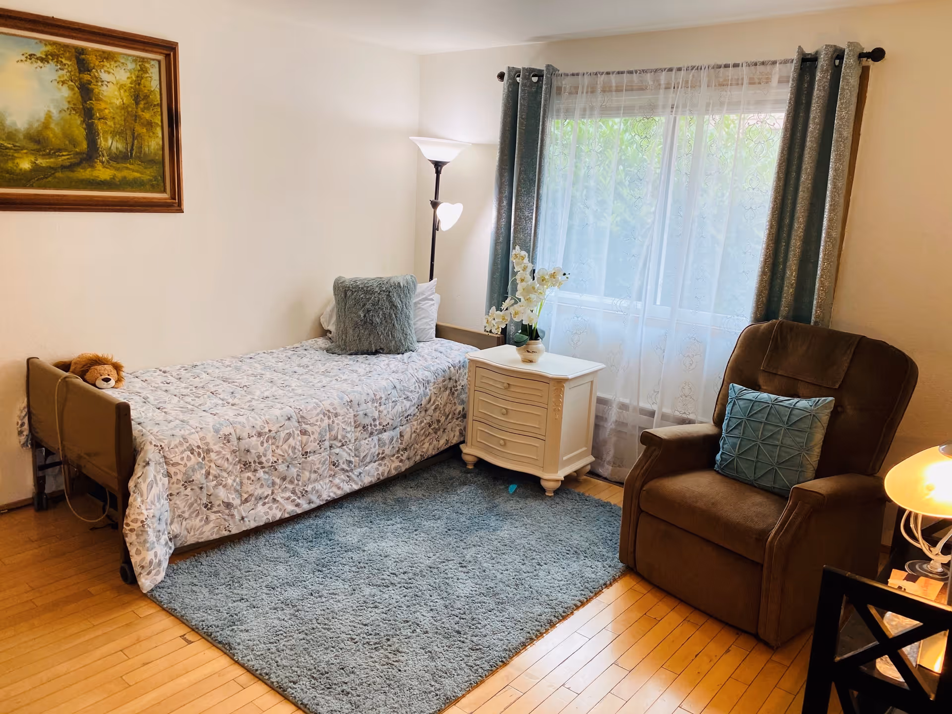 A cozy bedroom with a single bed covered in a patterned quilt and a plush pillow. A small stuffed lion toy rests on the bed. Next to the bed is a white nightstand with a vase of white flowers. A tall floor lamp stands behind the nightstand. A large window with sheer white curtains and blue drapes lets in natural light. A brown armchair with a blue cushion is positioned near the window. The floor is wooden with a blue area rug in front of the bed.