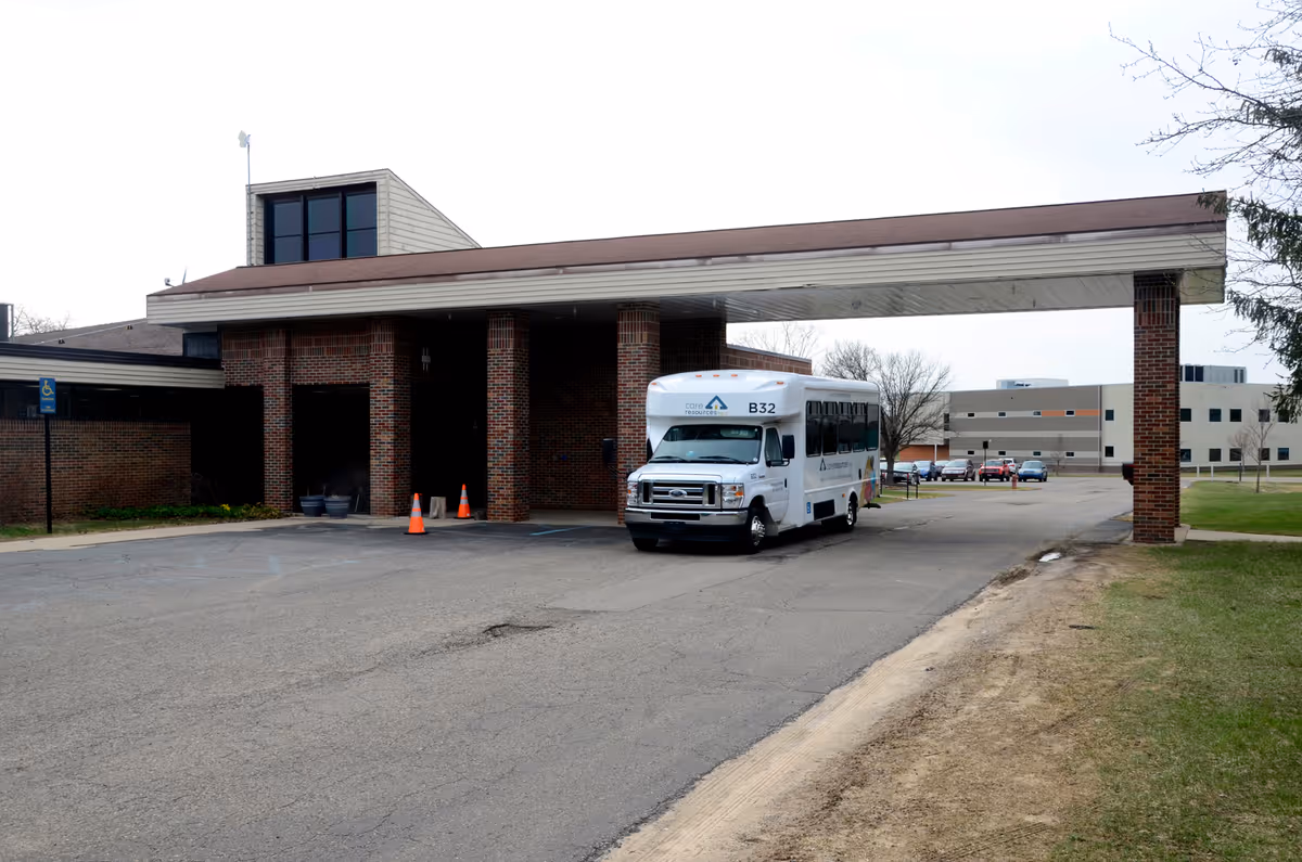 A white bus pulls up to a brick care facility under a covered driveway.