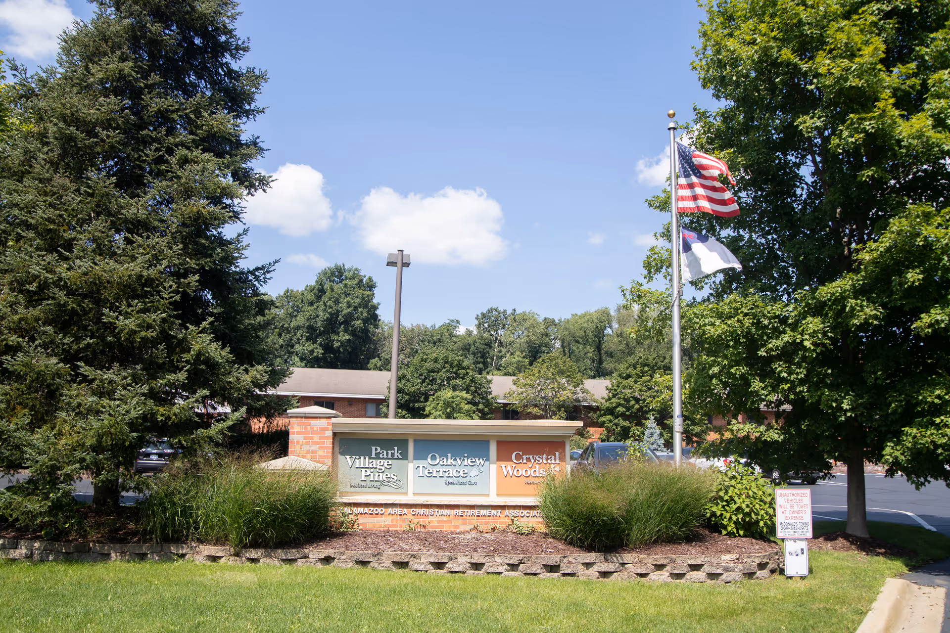 Entrance sign reading "Park Village Pines" with flags, landscaping, and a low brick building in the background.