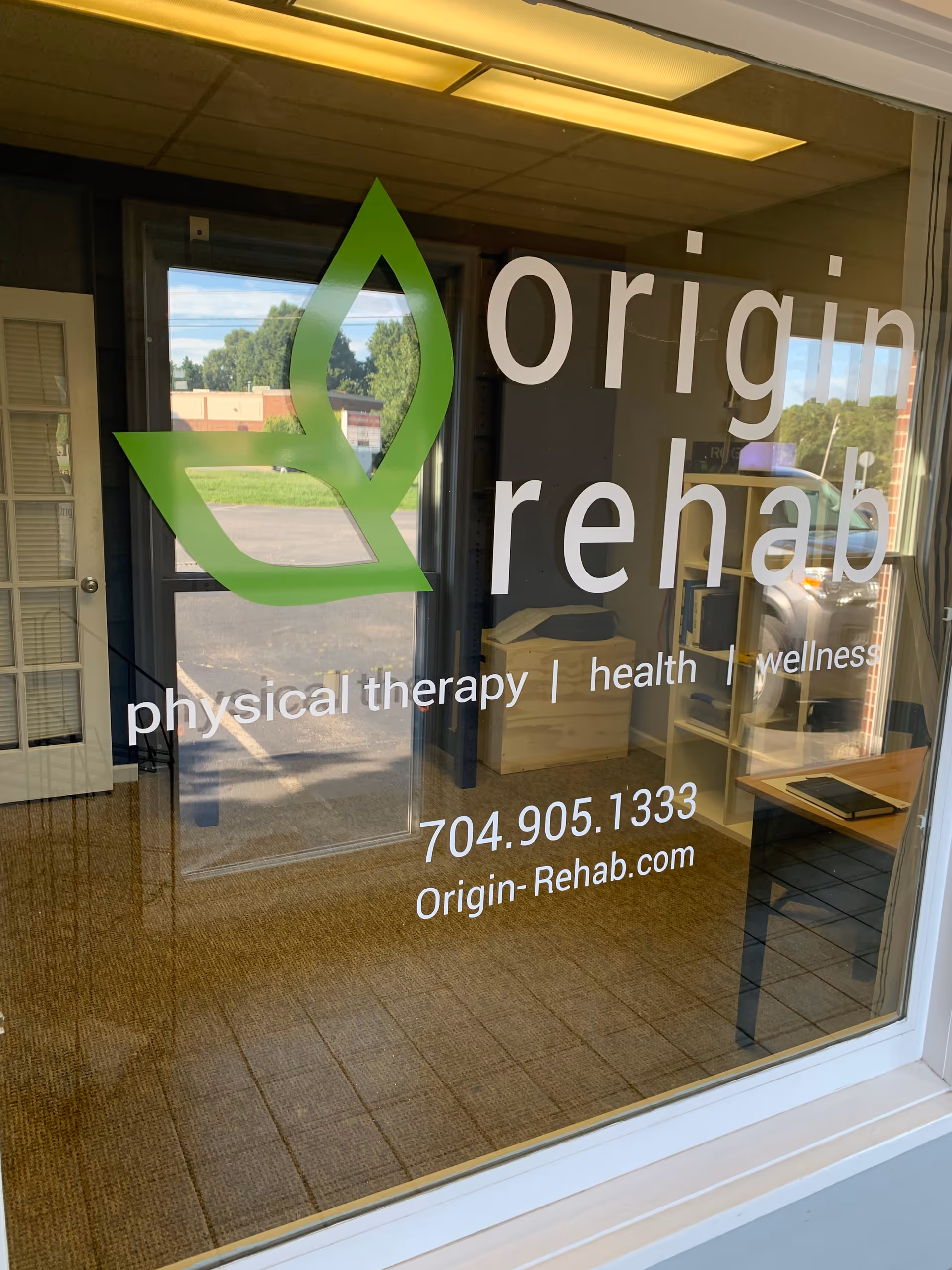 Storefront window displays Origin Rehab logo and signage for physical therapy services.
