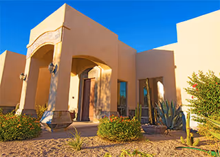 Stucco facility facade with arched entrance, desert landscaping, and a clear blue sky.