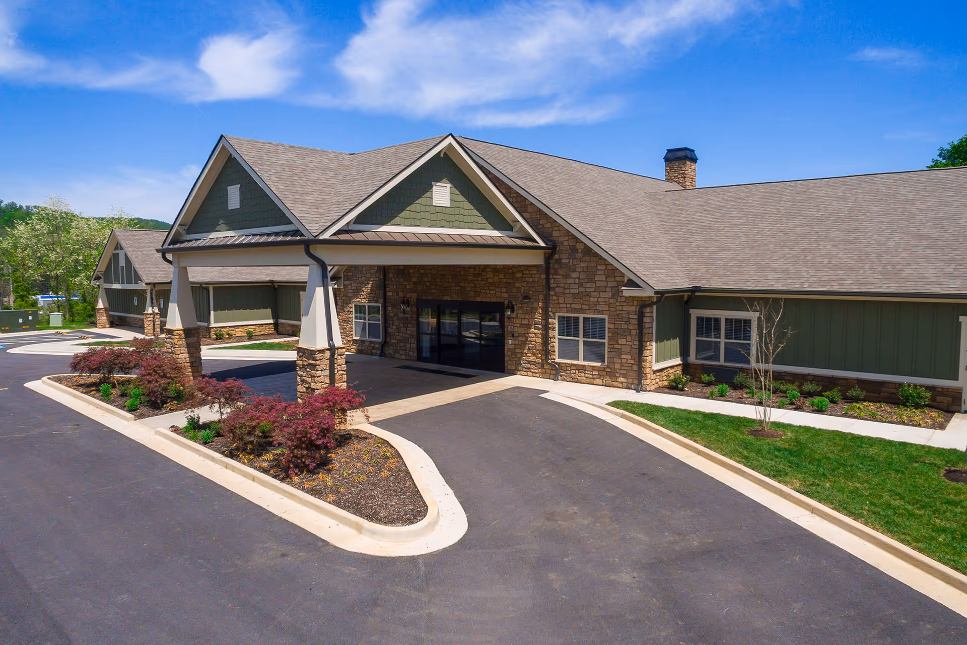 Front entrance of a single-story senior living facility with a covered drop-off, stone façade, green siding, and landscaped drive.