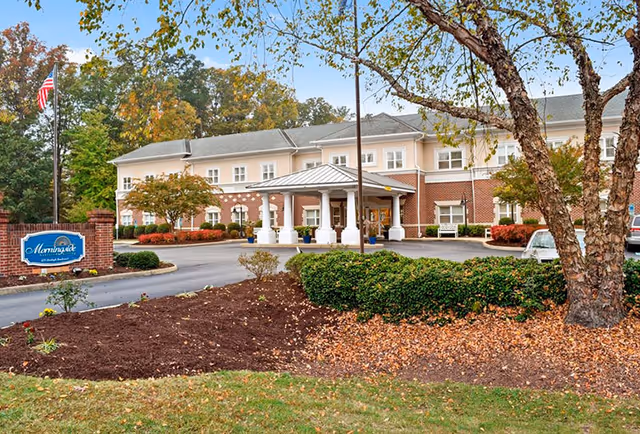 Exterior view of a two-story senior living facility with a covered entrance supported by white columns. The building has a brick lower facade and light-colored upper walls. There is a landscaped area with bushes and trees in front, and an American flag on a flagpole near the entrance. A blue sign with white text is visible on the left side of the driveway.