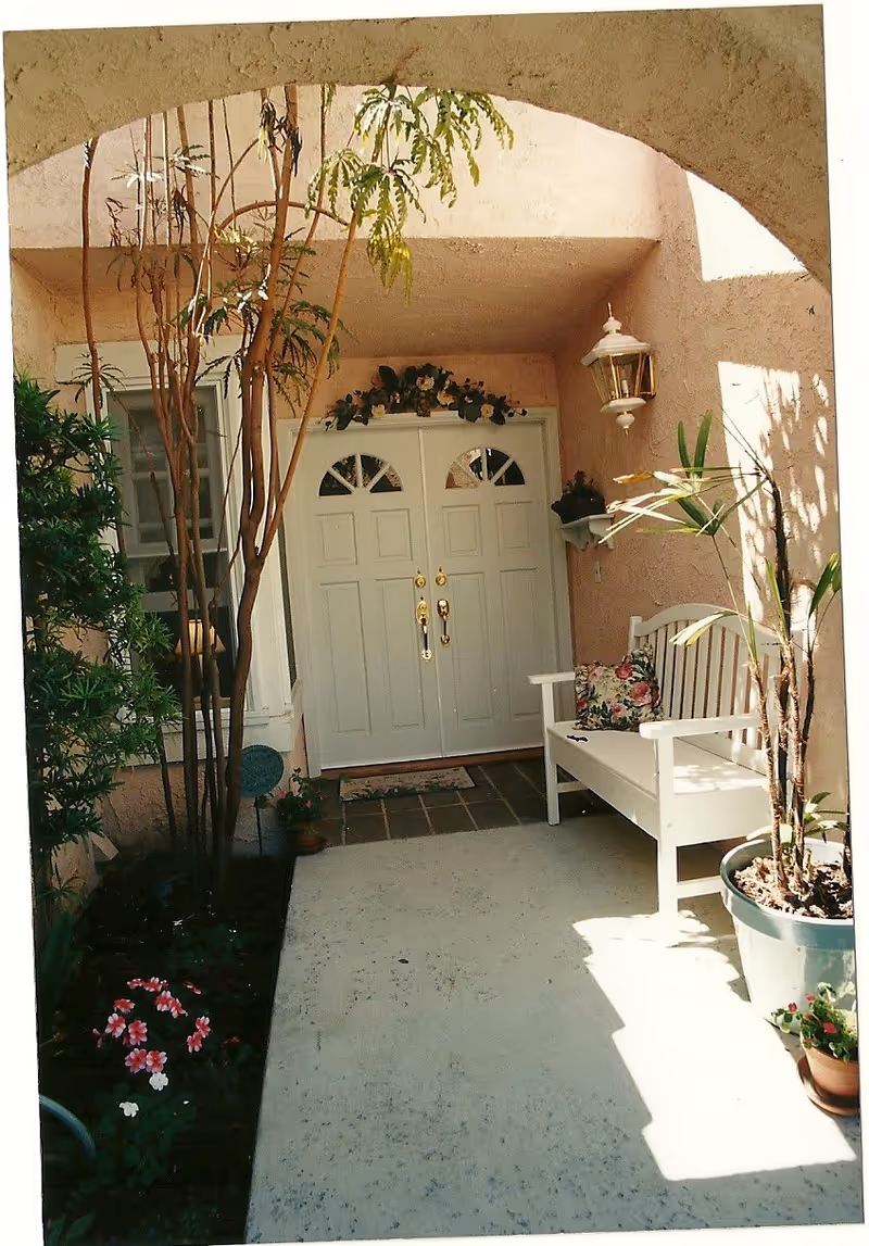 Covered front porch with double white entry doors, a white bench, potted plants and an arched stucco entryway.