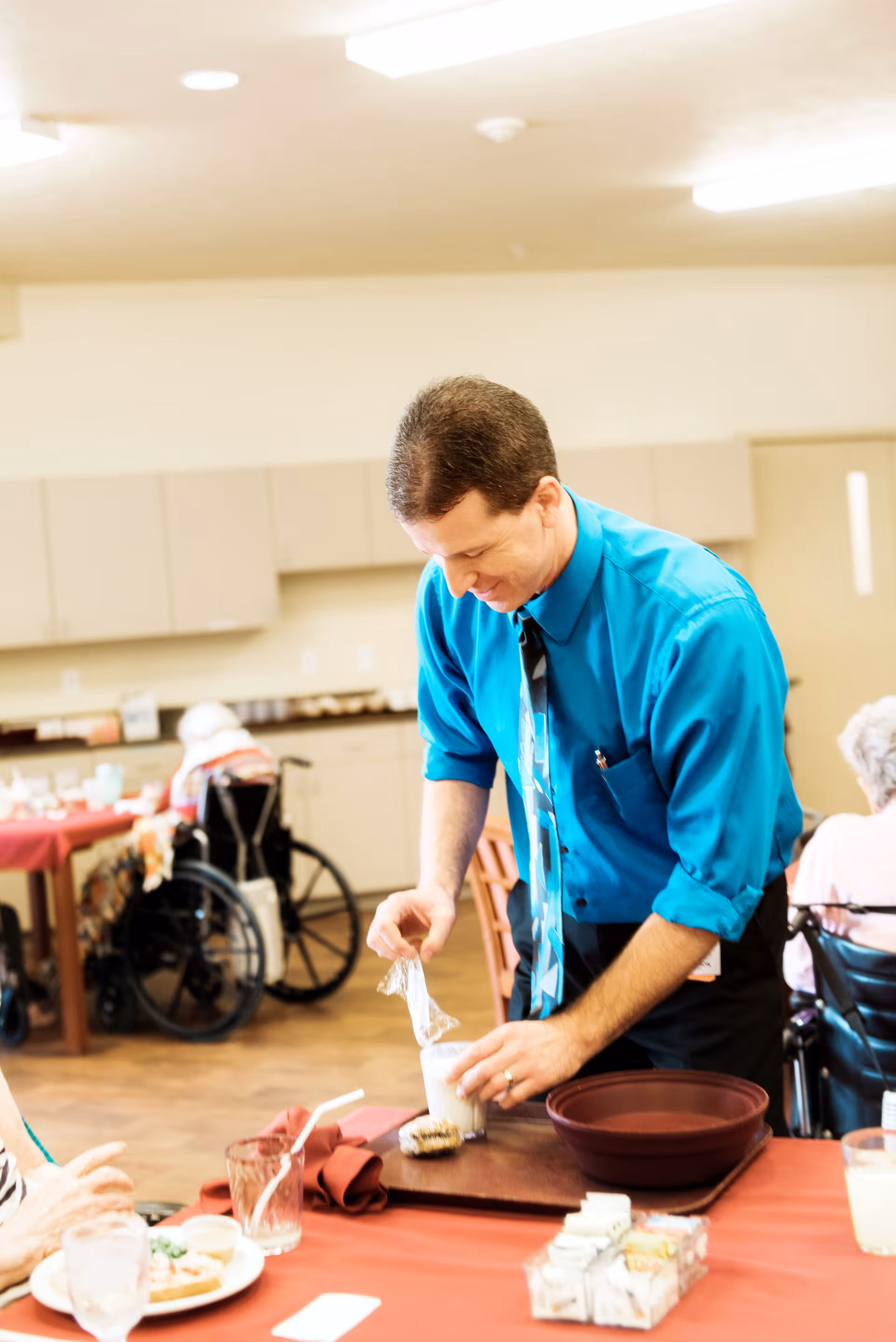 A staff member in a teal shirt serves a dessert at a dining table in a senior care facility dining room with residents and wheelchairs in the background.
