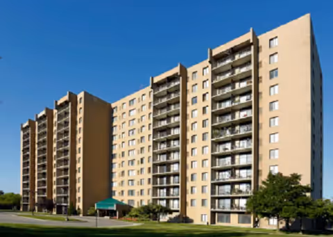 Tall beige apartment complex with balconies and a green entrance canopy under clear blue sky.