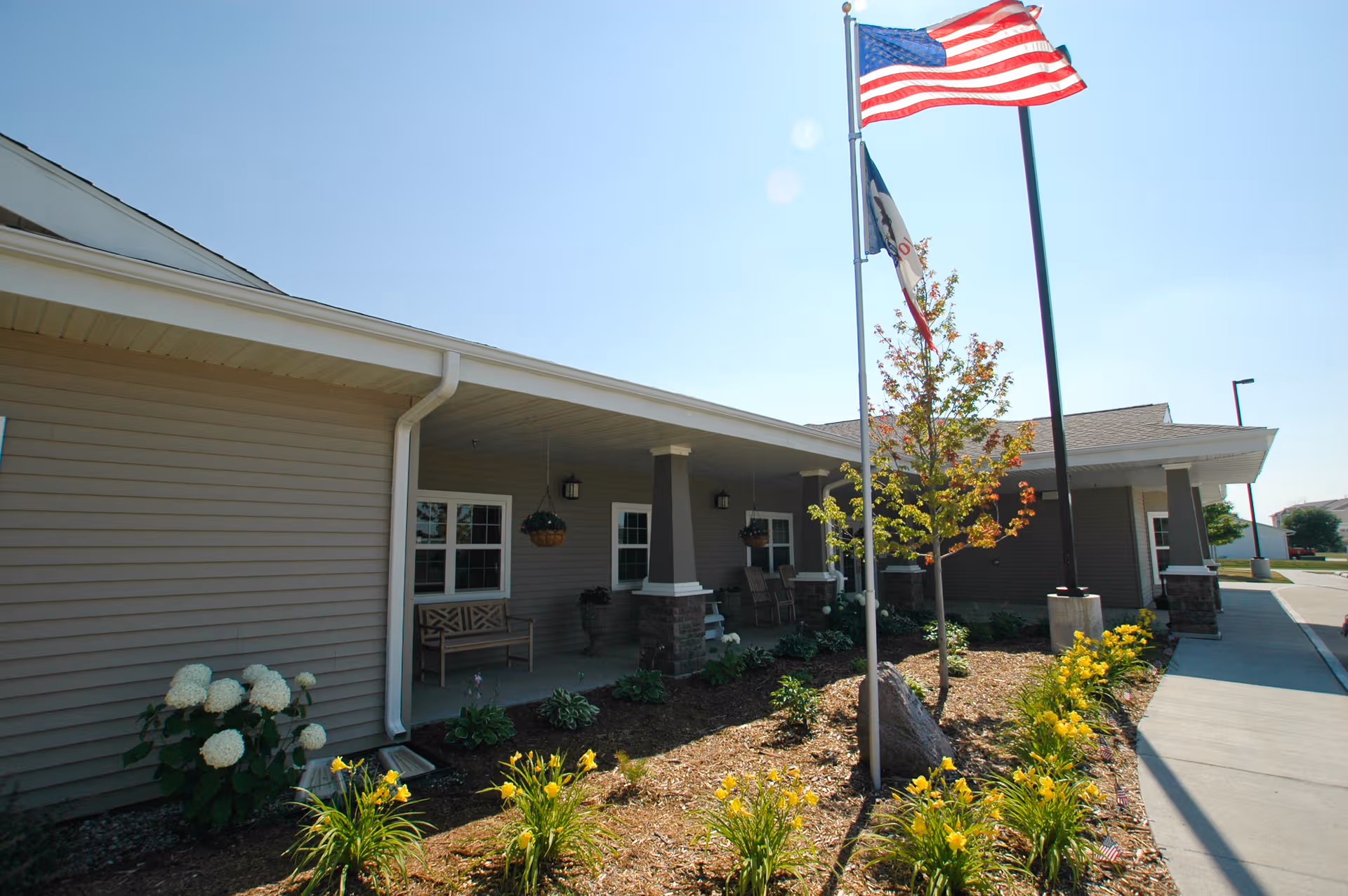 Single-story senior living facility exterior with a covered porch, flags, and landscaped front garden.