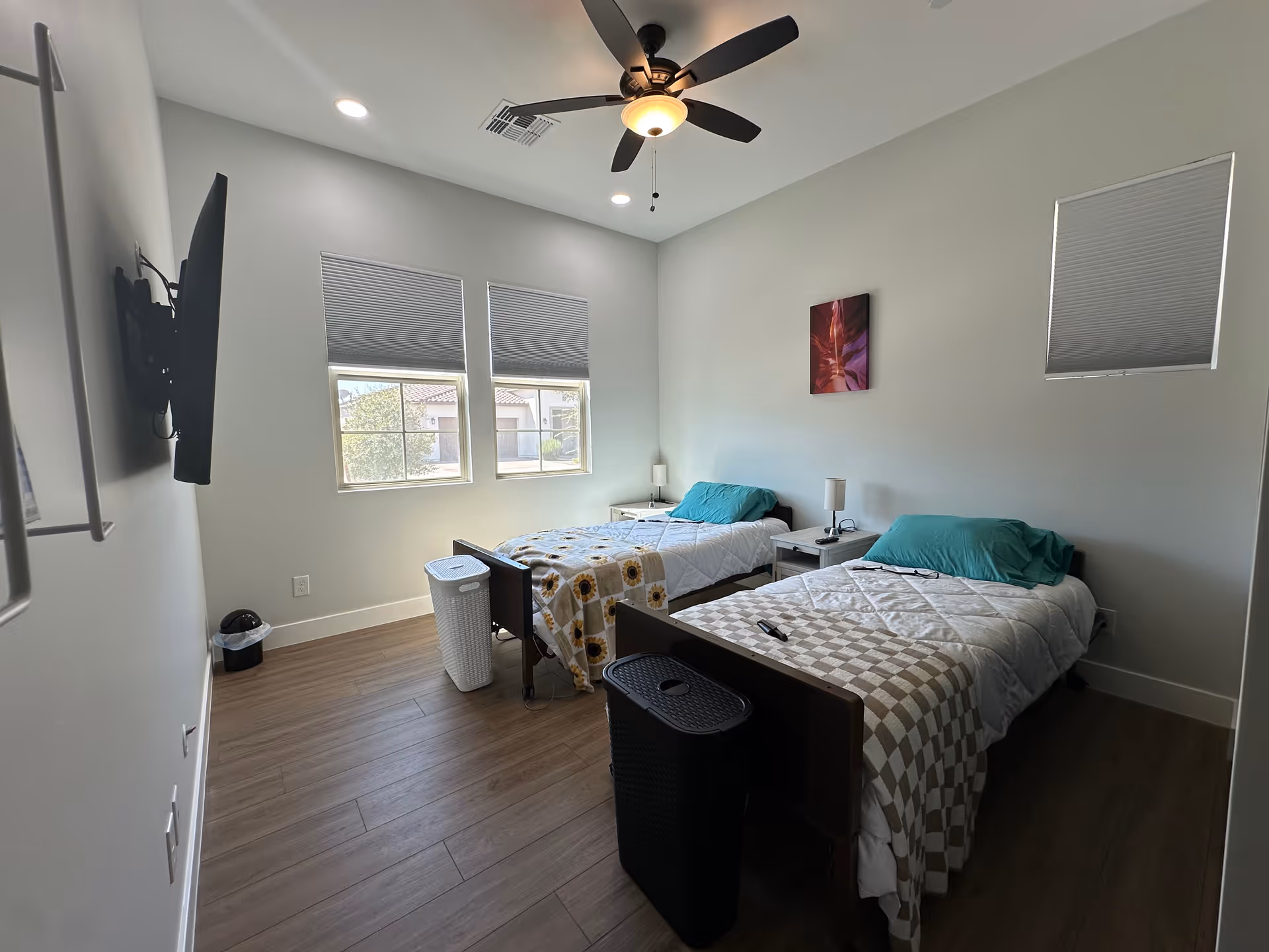 Bright shared bedroom with two twin beds, teal pillows, ceiling fan, and windows.