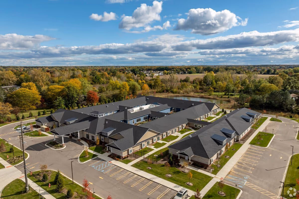 Aerial view of a single-story senior living complex with connected buildings, parking lots, and surrounding trees and fields.