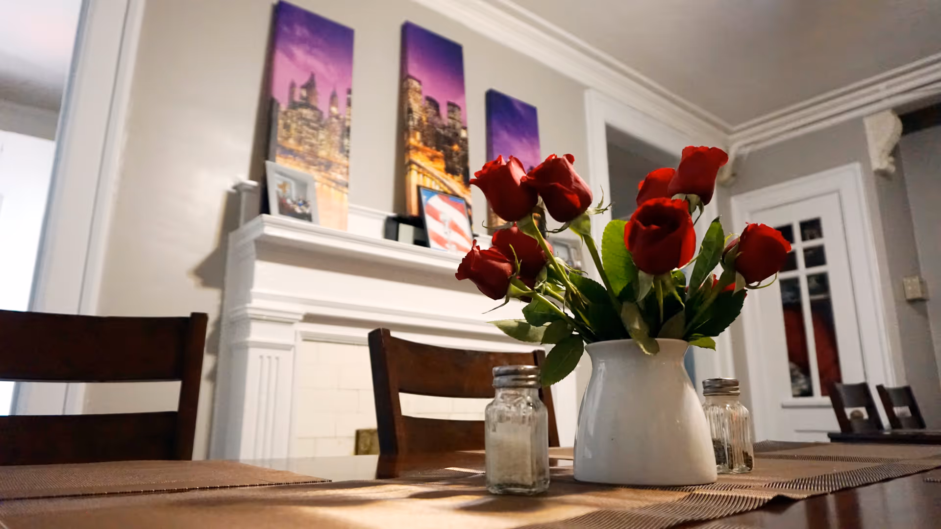 A dining table with a white vase holding red roses and salt and pepper shakers. In the background, there is a white fireplace mantel with framed pictures and three vertical cityscape paintings with purple skies above it. Wooden chairs surround the table, and a door with glass panes is visible on the right side of the room.