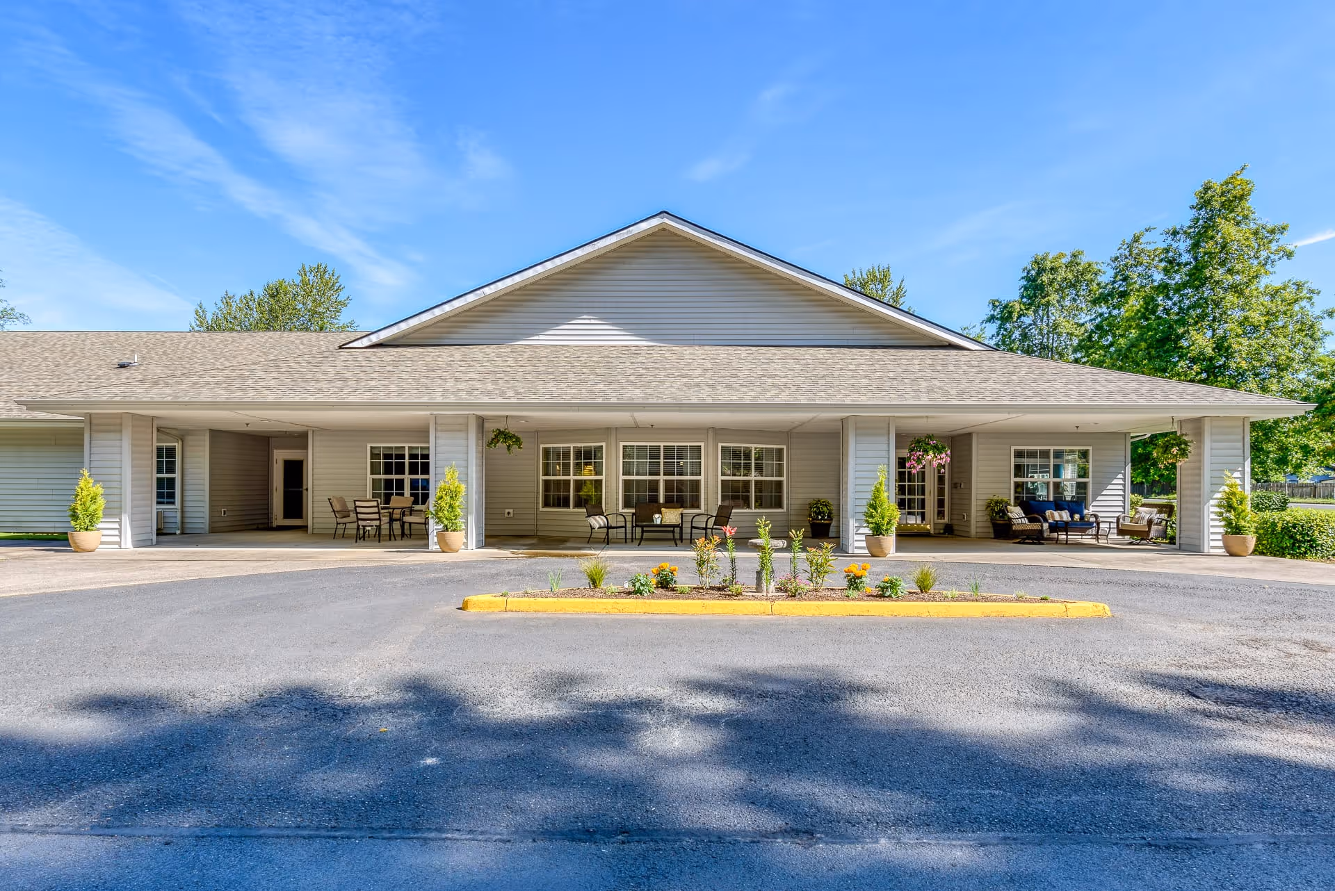 Exterior view of Creekside Place Memory Care building with a covered entrance area featuring outdoor seating, potted plants, and a small landscaped island in front of the driveway under a clear blue sky.