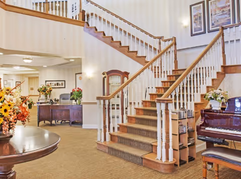 Elegant lobby with a grand staircase, piano, and floral arrangements in a senior living facility.