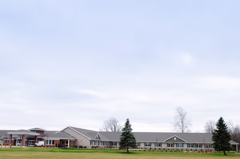 Wide exterior view of a single-story senior living facility building with a large grassy lawn in front, a few evergreen trees, and a cloudy sky overhead.