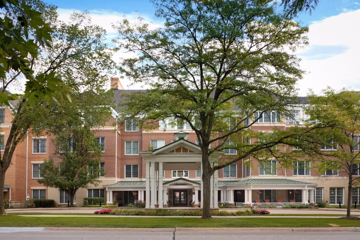 Front exterior of a multi-story red-brick senior living building with a covered portico entrance and large trees on the lawn.