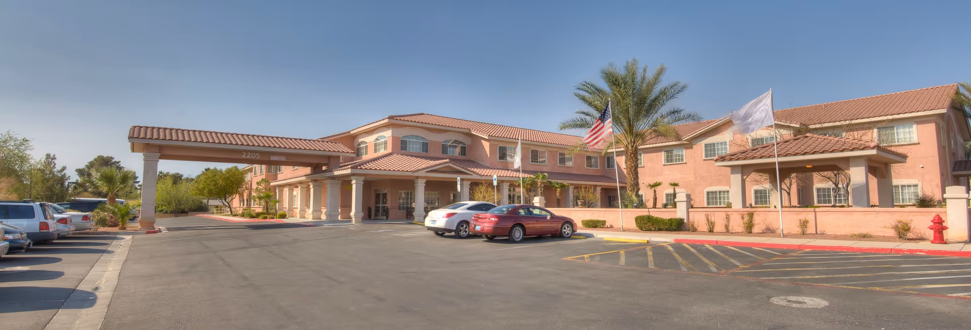Exterior view of The Bridge at Paradise Valley senior living facility showing a large two-story building with a covered entrance, several parked cars, palm trees, and flags including an American flag under a clear blue sky.