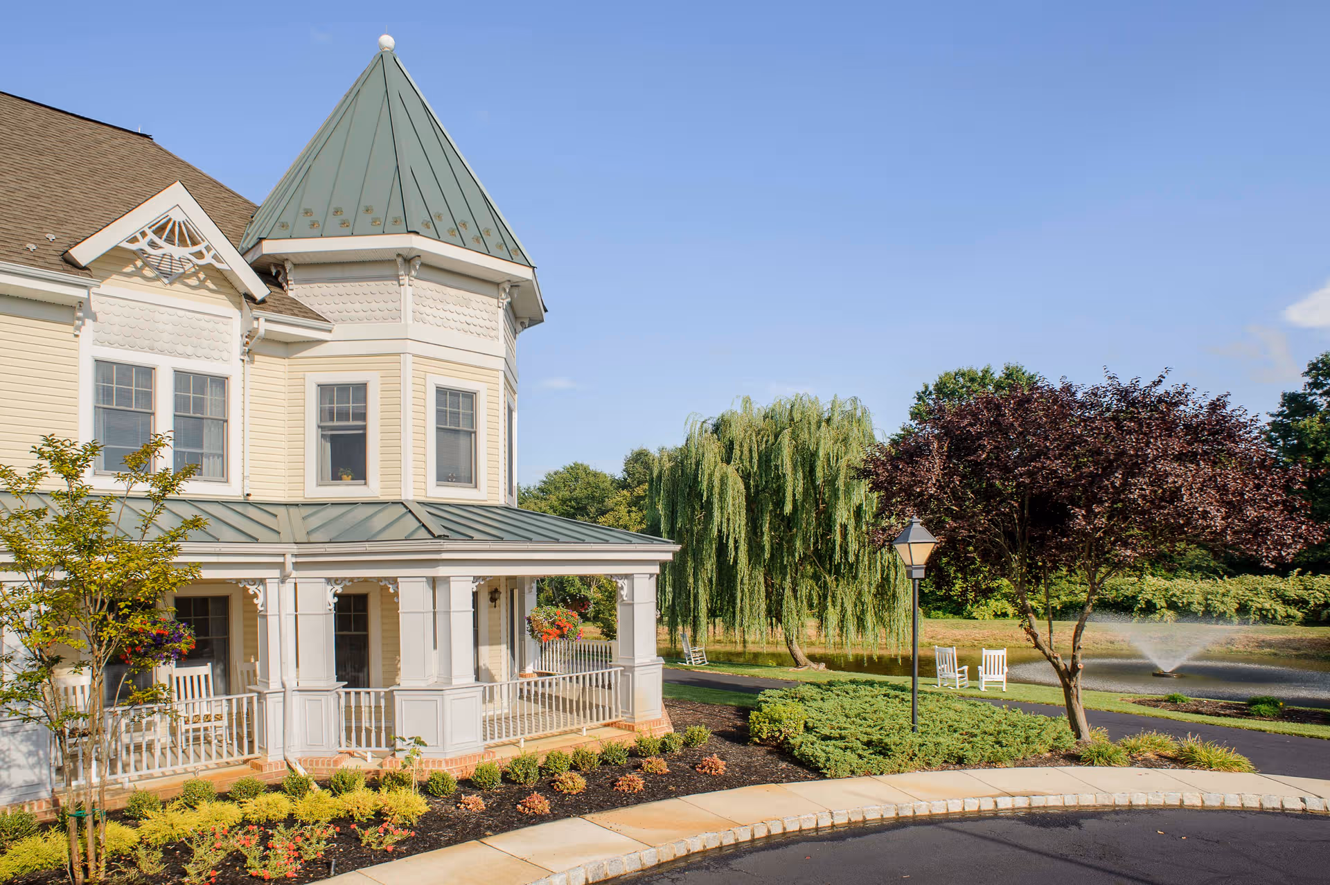 A light-colored, two-story Victorian-style senior living building with a turret, covered porch, and landscaped grounds.