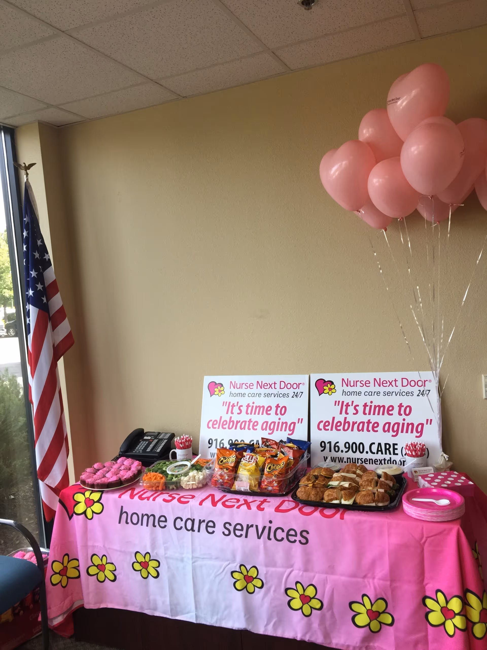 Indoor promotional table with pink balloons, decor, snacks, and Nurse Next Door signage.