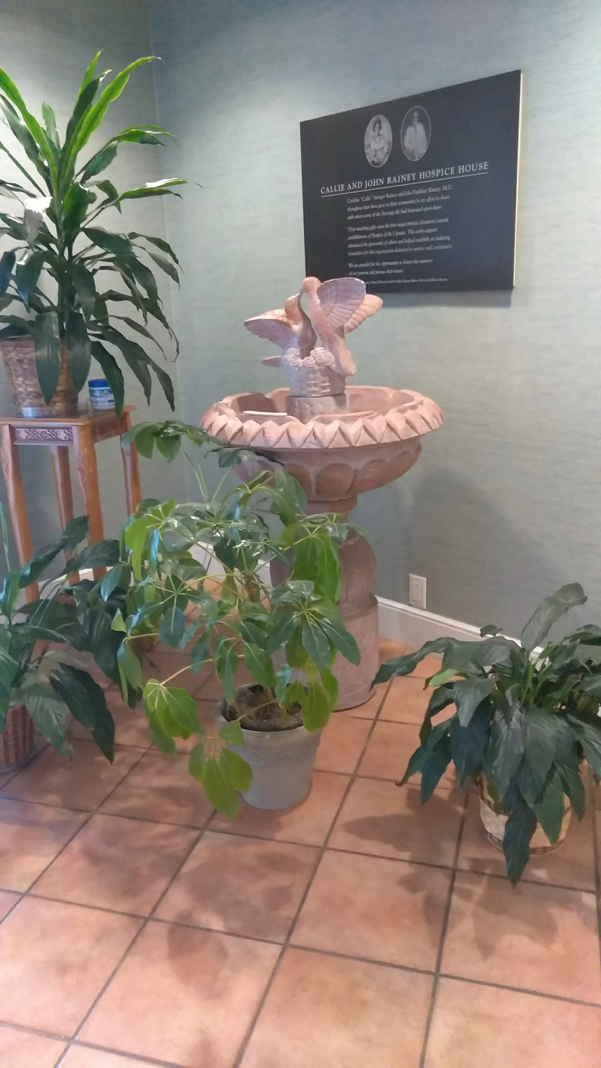 Indoor hospice lobby features potted plants, a decorative fountain, and a wall plaque.