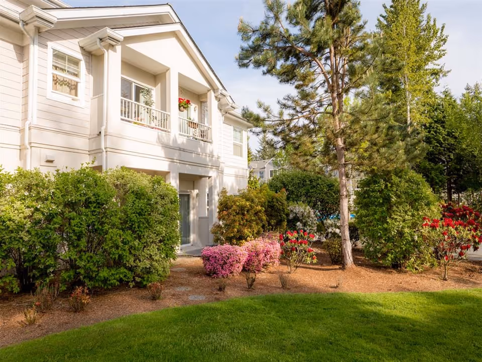 Exterior view of a light-colored two-story building with a balcony, landscaped shrubs, flowering plants, and trees.