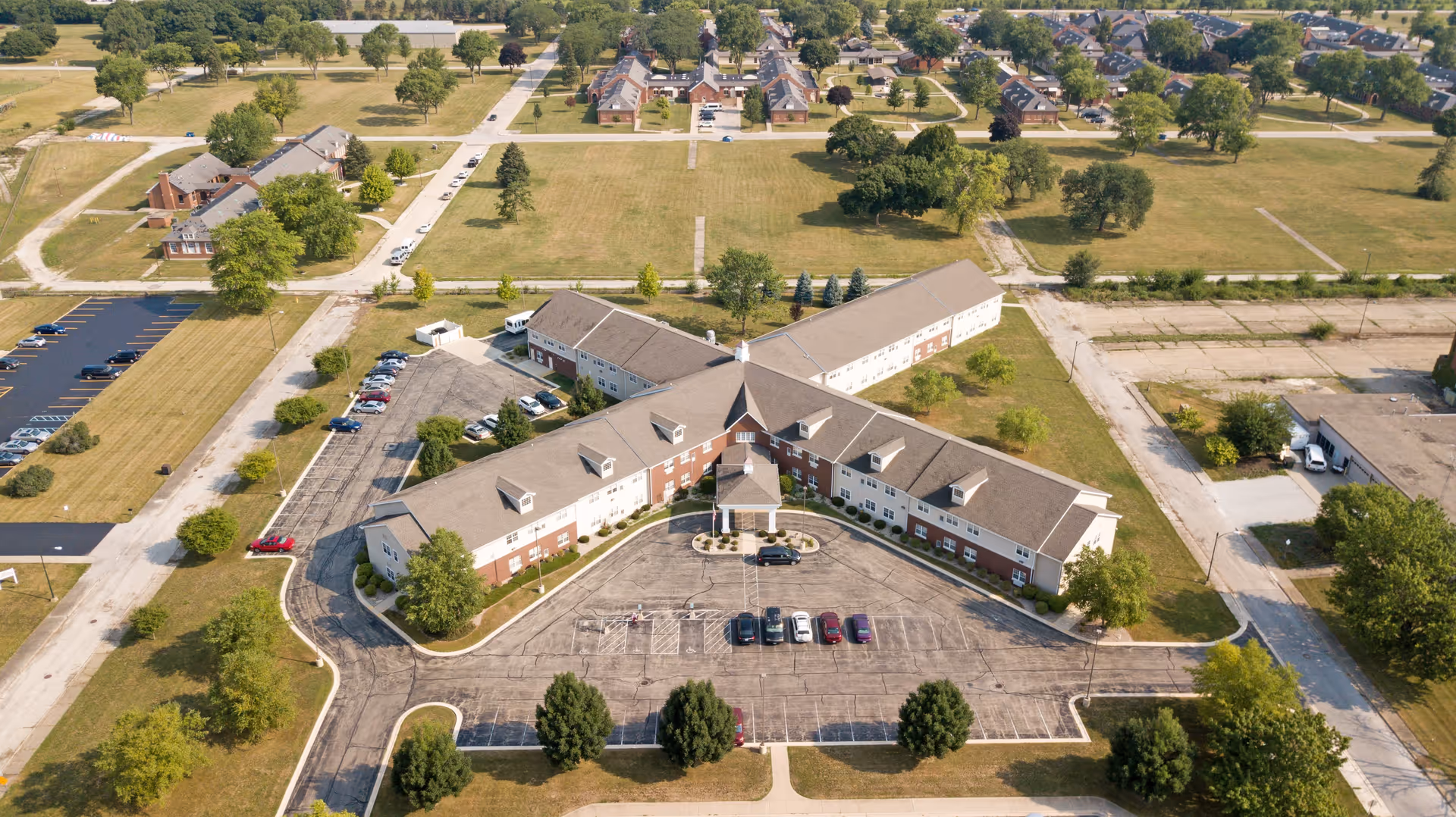 Aerial view of Heritage Woods of Manteno senior living facility showing a large, two-story building with a triangular shape surrounded by parking lots and green lawns. Several cars are parked in front of the main entrance, and there are trees and open grassy areas around the property. Residential houses and streets are visible in the background.