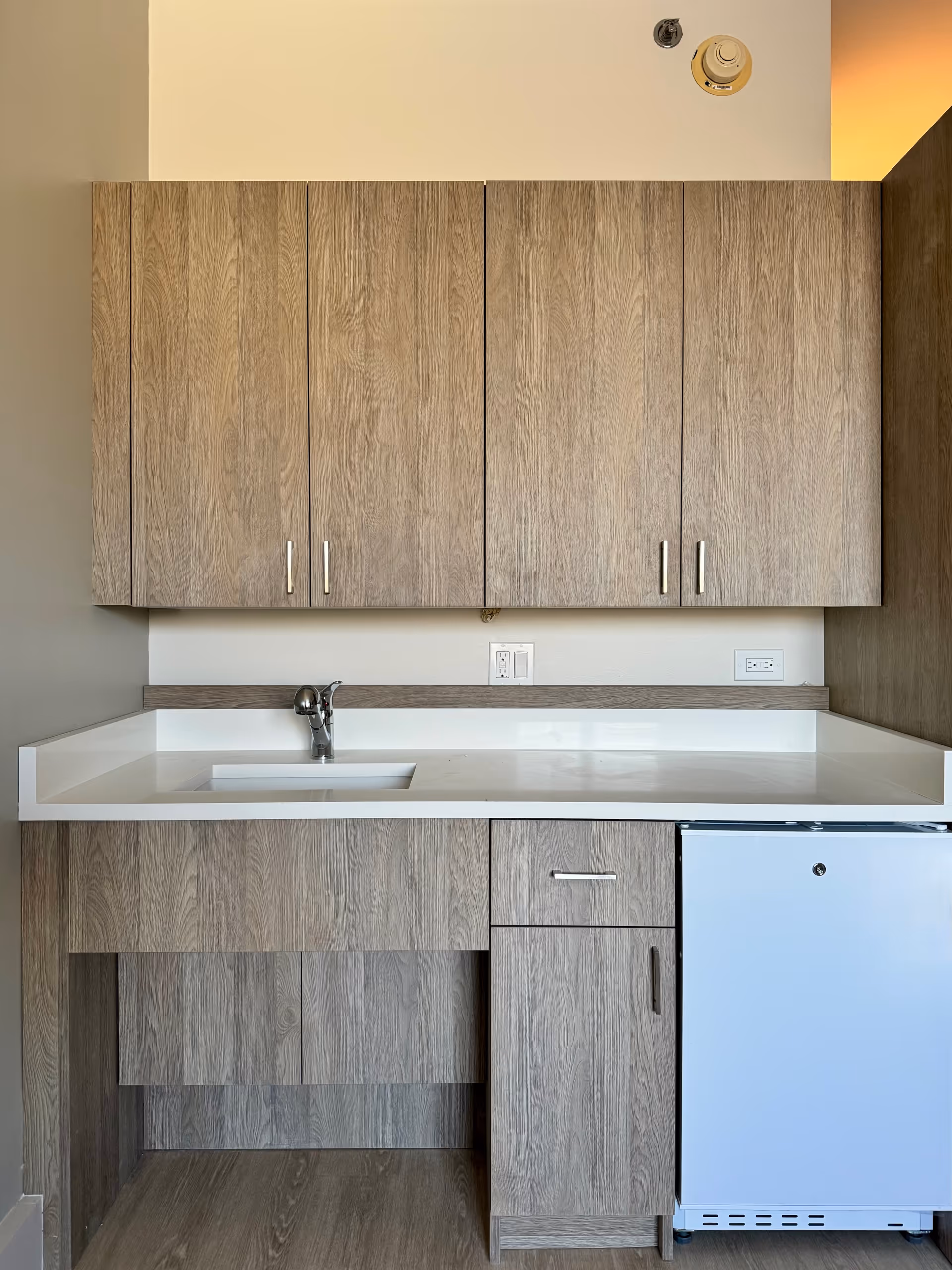 Wooden cabinets line a kitchenette with sink, countertop, and small refrigerator.
