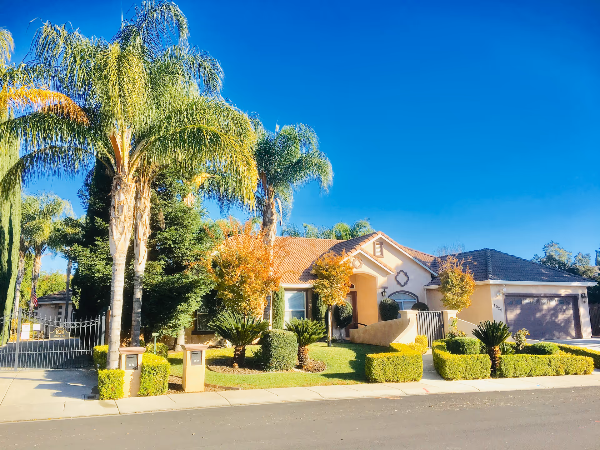 Exterior view of a single-story residential building with a tiled roof, surrounded by well-maintained landscaping including palm trees, bushes, and small trees under a clear blue sky.