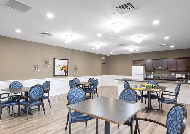 Well-lit dining/activity room with multiple tables and blue-upholstered chairs and a kitchenette along the back wall.
