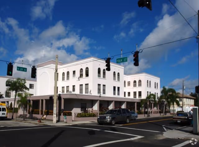 Exterior view of a three-story white building at the corner of N 20 Ave and Polk St under a partly cloudy blue sky. Several cars are parked along the street and palm trees are visible around the building.
