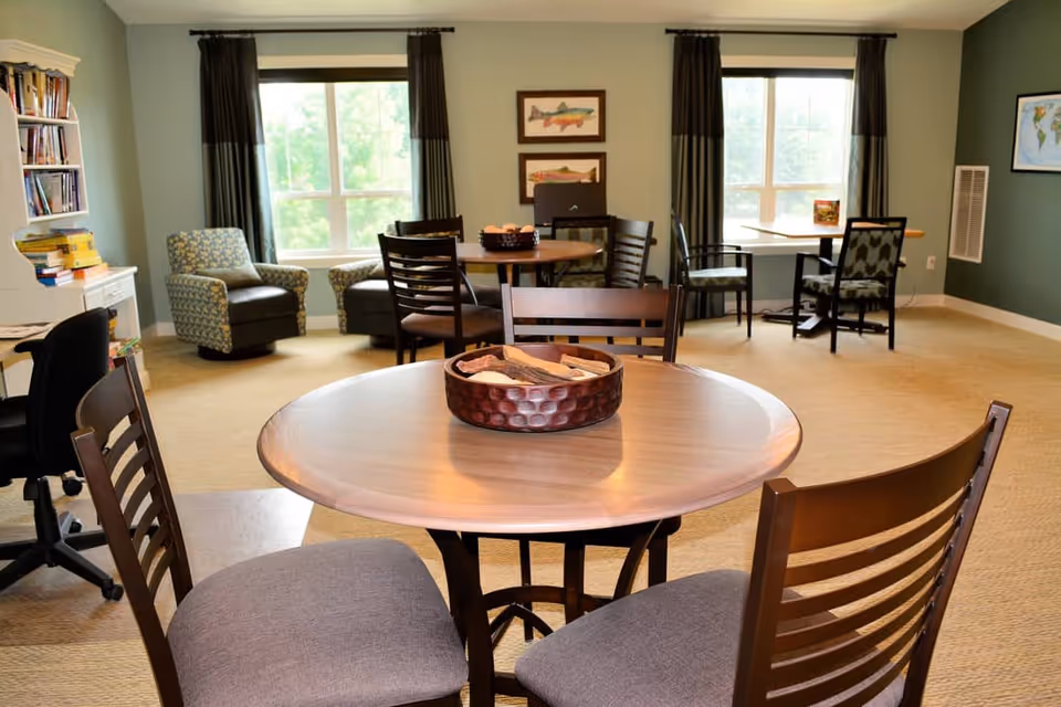 A cozy common area in a senior living facility with round wooden tables and cushioned chairs. The room has large windows with dark curtains letting in natural light, two patterned armchairs near a bookshelf filled with books, and framed fish artwork on the wall. The carpeted floor and soft green walls create a warm and inviting atmosphere.