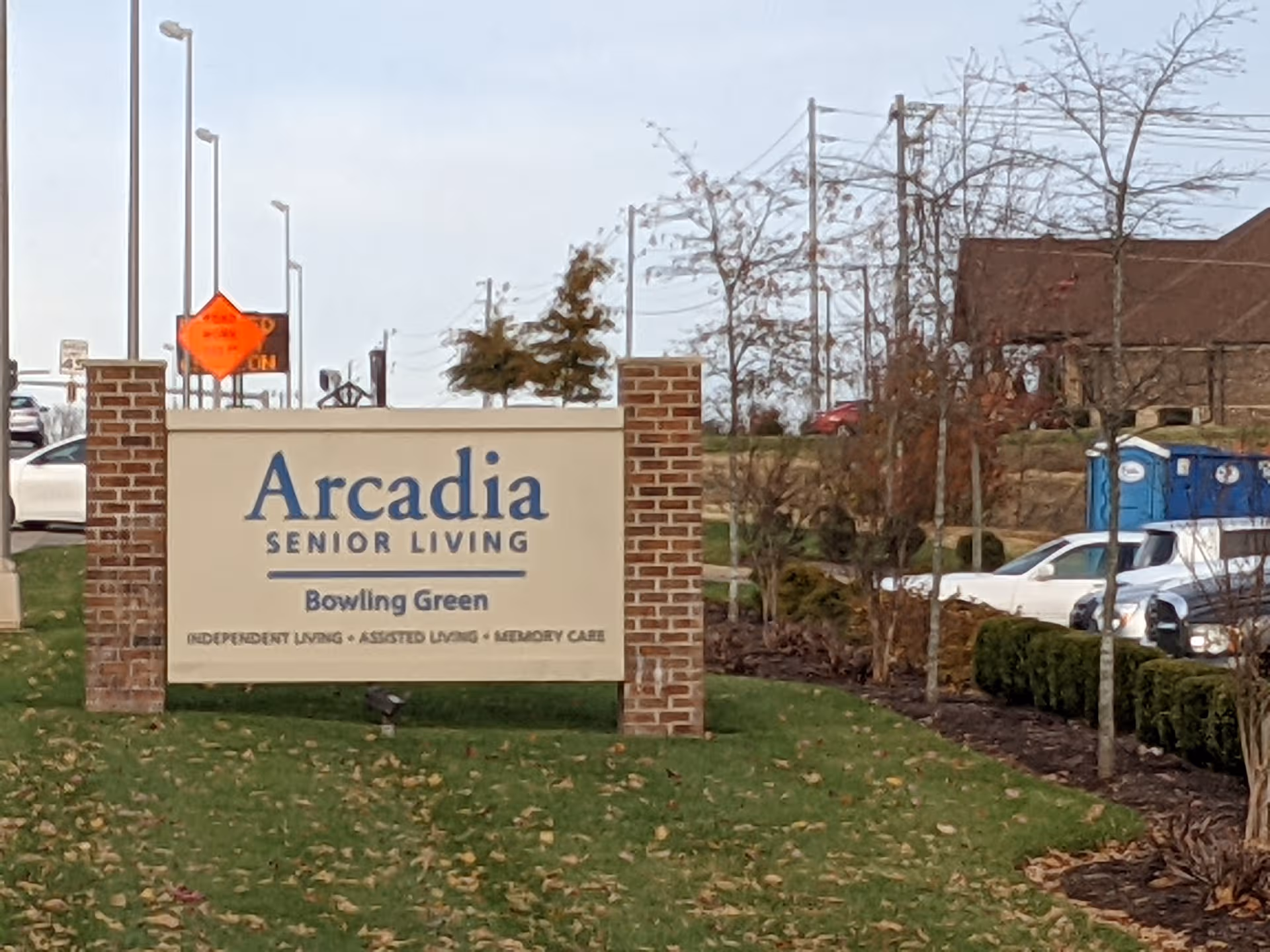 Arcadia Senior Living sign with Bowling Green, brick pillars, shrubs, and parked cars.