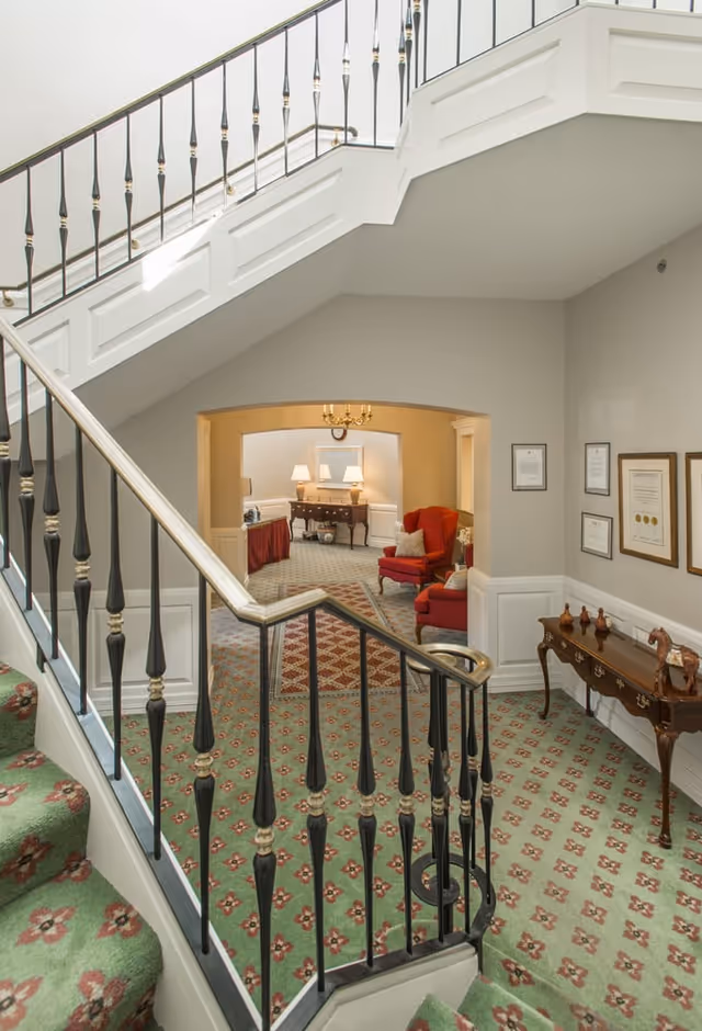 Elegant staircase inside a senior living facility lobby, with patterned carpet and red seating beyond.