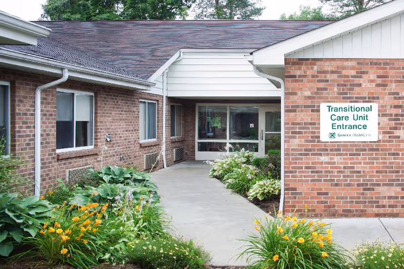 Brick building front with a walkway, landscaped flower beds, and a sign reading 'Transitional Care Unit Entrance'.