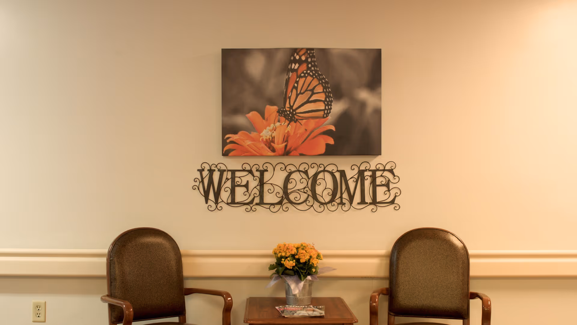 Interior lobby with butterfly artwork, a decorative 'Welcome' sign, a small table, and two brown chairs.