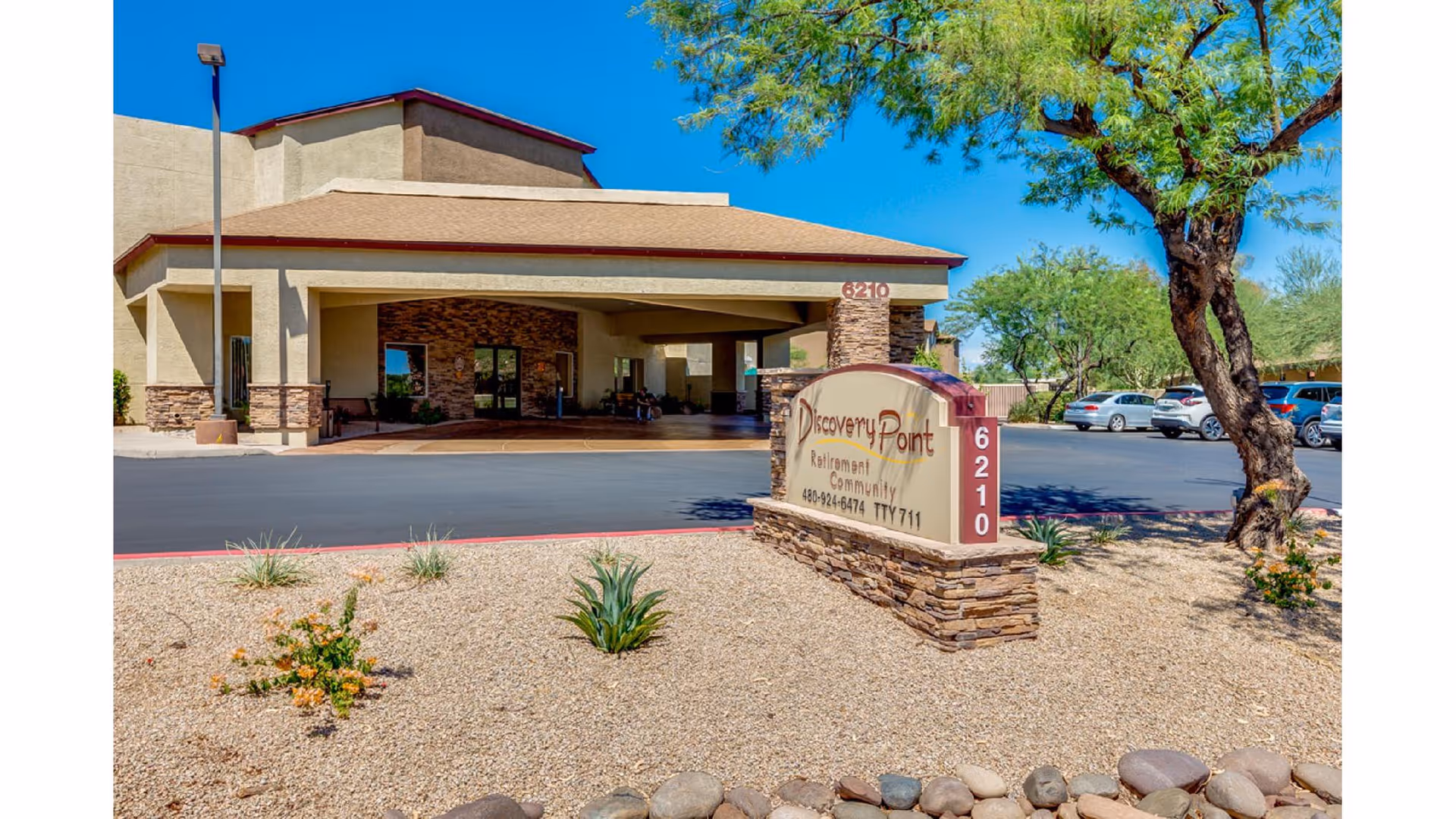 Exterior view of Discovery Point Retirement Community building entrance with a covered drop-off area, desert landscaping with rocks and plants, a tree, and a sign displaying the facility name and address 6210. Several parked cars are visible in the background under a clear blue sky.
