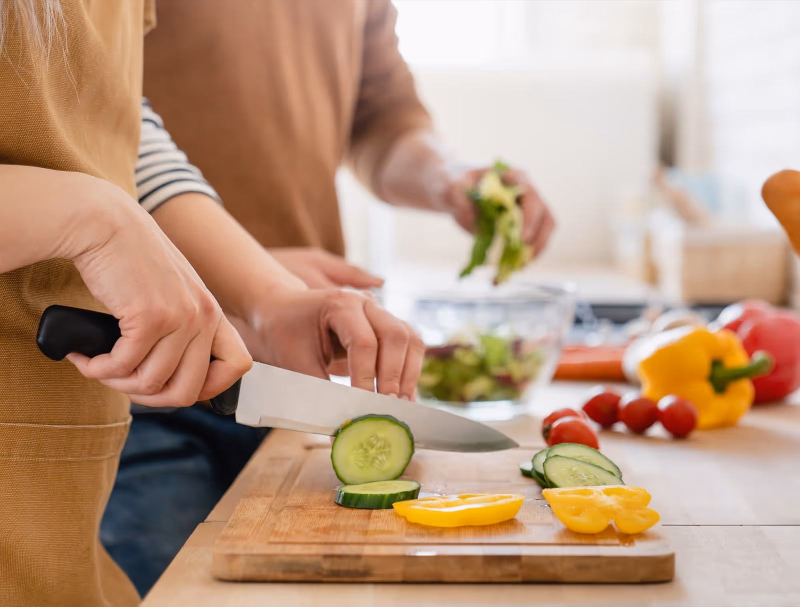 Two people slice cucumbers on a cutting board in a bright kitchen.