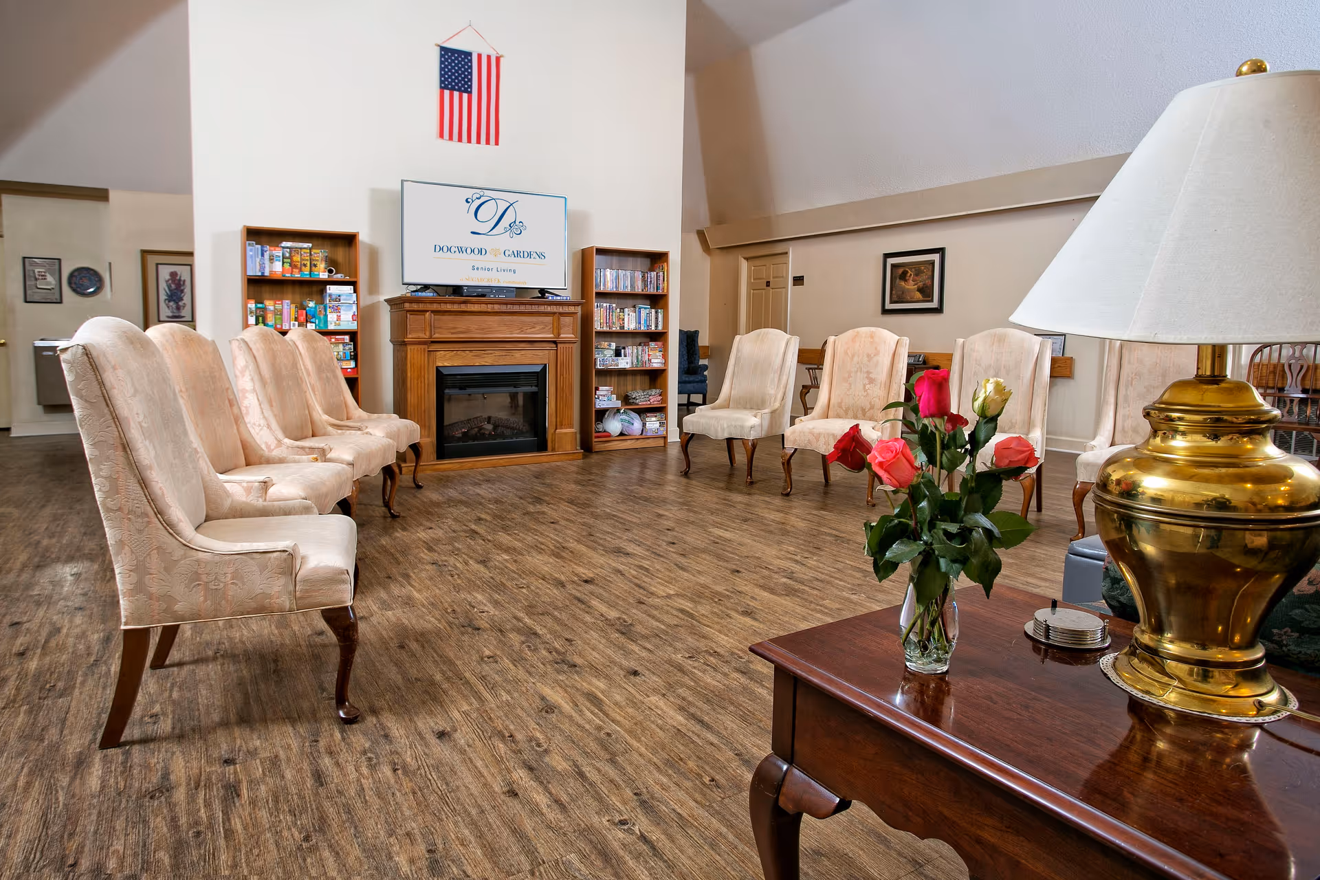 A senior living facility common area with a semi-circle arrangement of beige upholstered chairs around a wooden fireplace with a TV above it displaying the Dogwood Gardens Senior Living logo. There are bookshelves with games and DVDs on either side of the fireplace, an American flag hanging on the wall above, and a wooden table with a brass lamp and a vase of roses in the foreground.