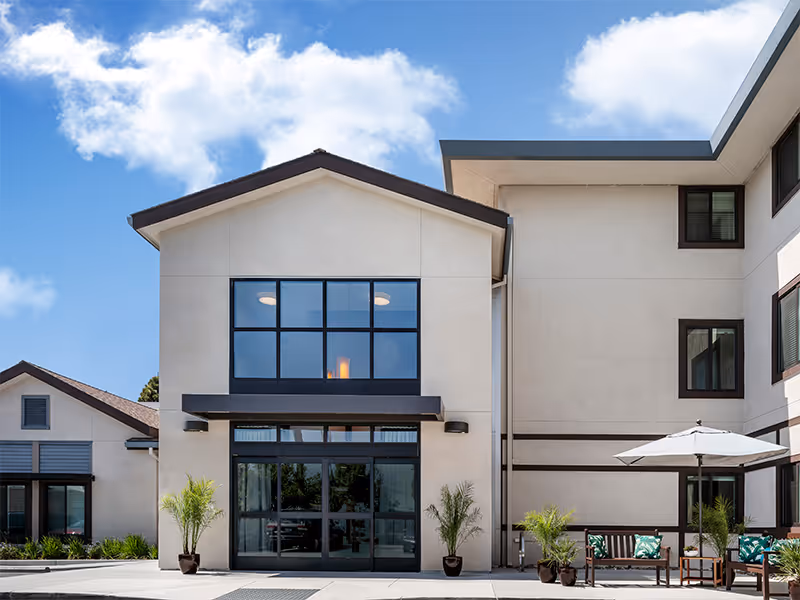 Modern senior living facility exterior with large glass entrance and outdoor seating.