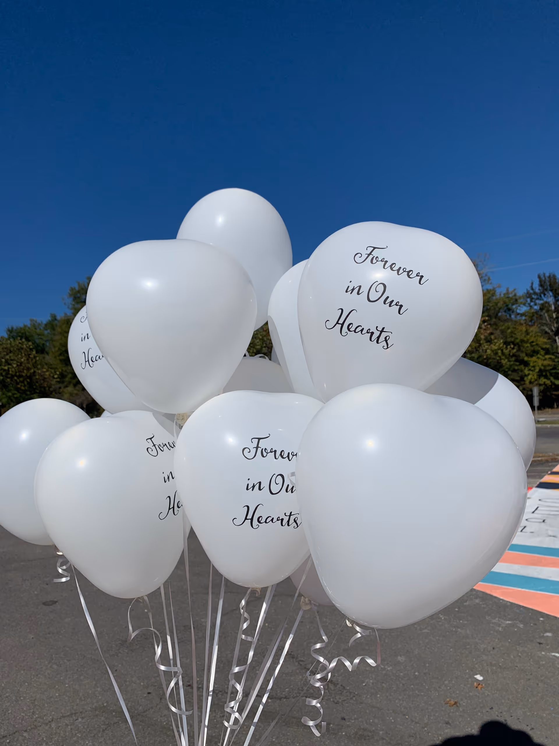 Cluster of white heart-shaped balloons with 'Forever in Our Hearts' outdoors on pavement.