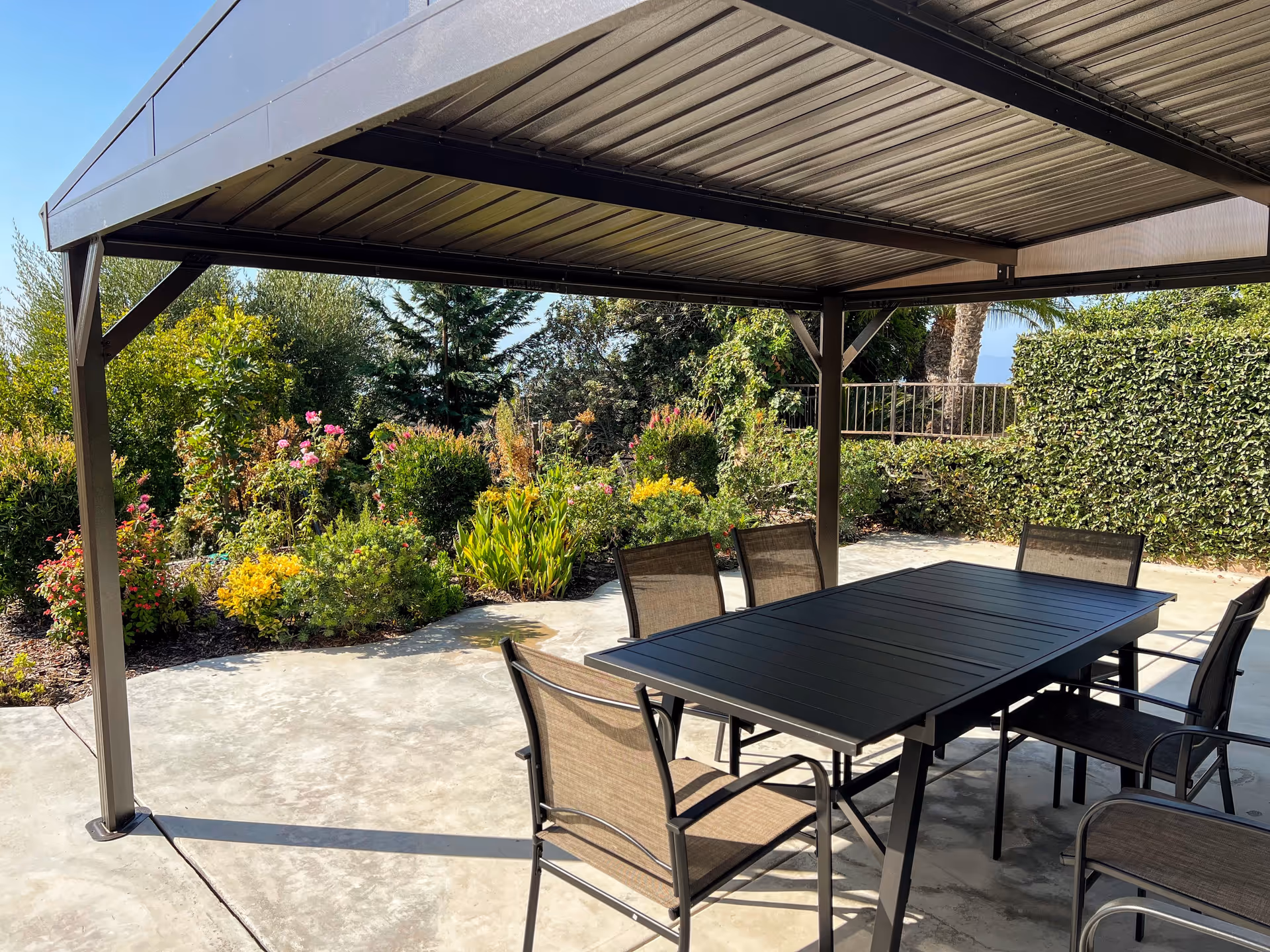 Outdoor patio under a metal pergola with a table, chairs, and lush garden surroundings.